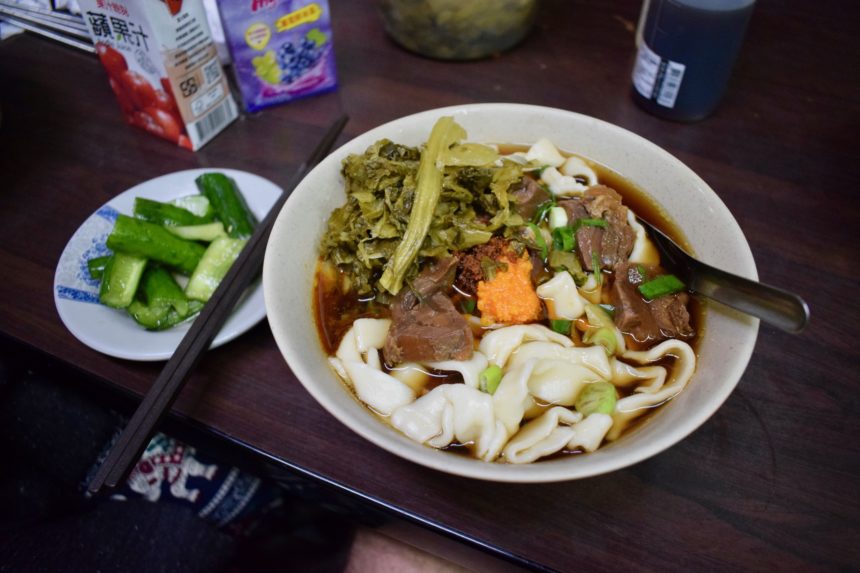 Bowl of best beef noodles in Ximending at Fuhong Beef Noodle with chilli butter and pickled mustard greens next to plate of pickled cucumbers