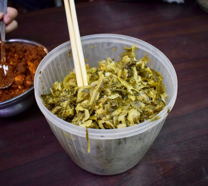 Large plastic pot filled with pickled mustard greens with a pair of chopsticks sticking out ready for self-serve