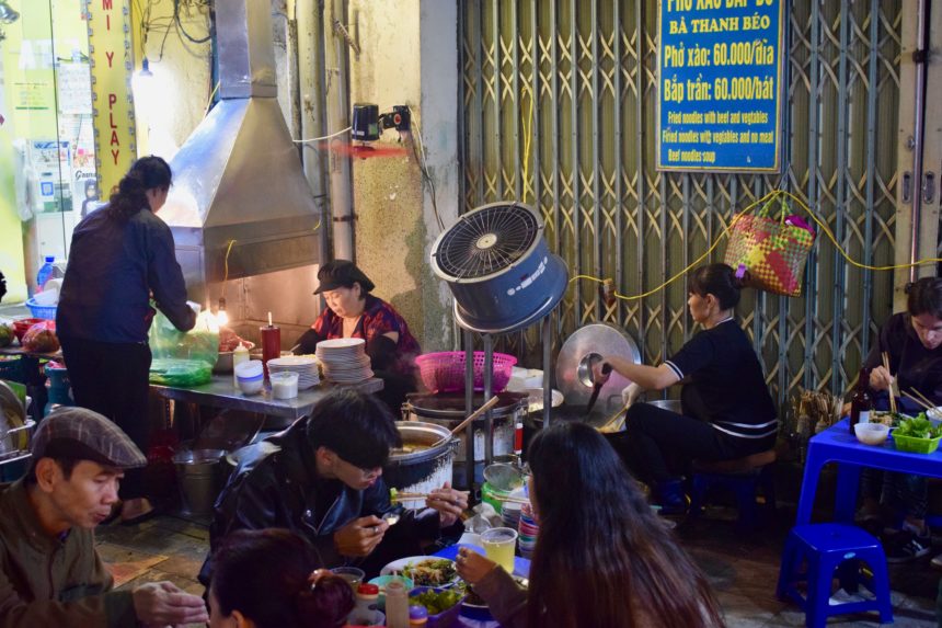 2 ladies cooking pho xao in hanoi in 2 separate woks being blown by a giant electric fan