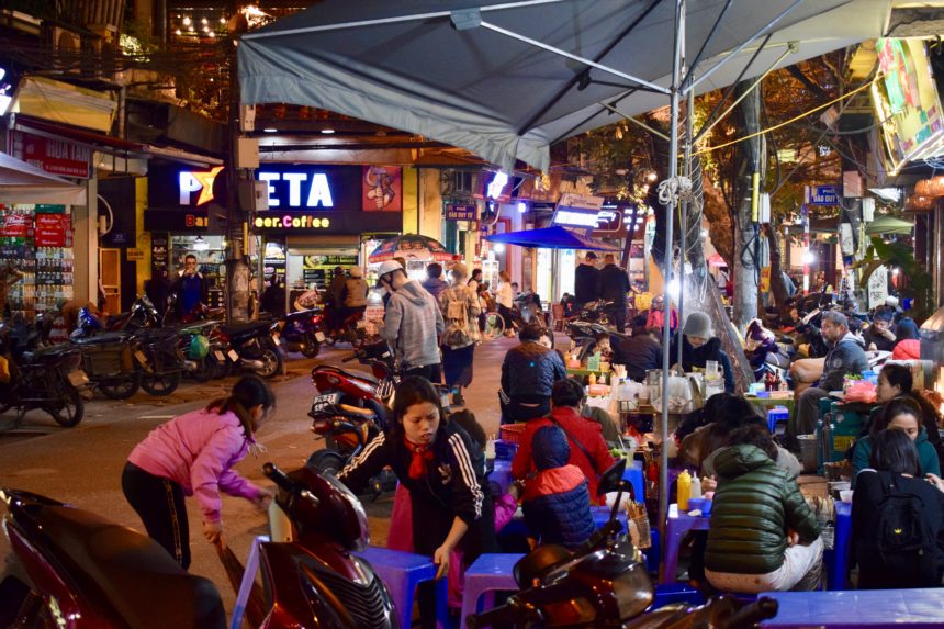 The busy Hanoi street with motorbikes passing next to the tables of Phở Xào Ba Thanh Béo