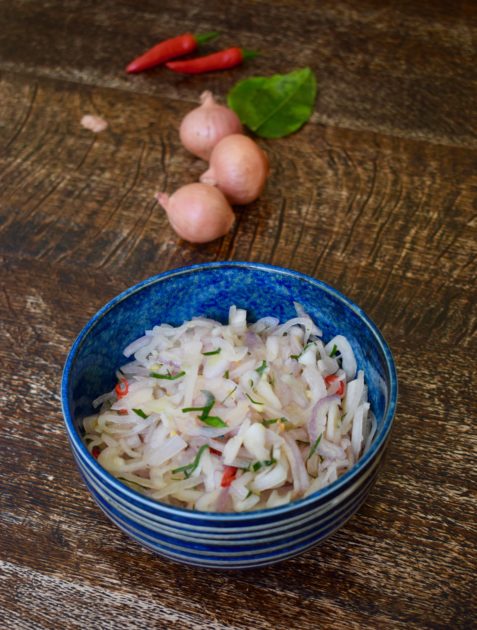 Raw sambal in a blue bowl with shallots kaffir lime leaves and chilli in background