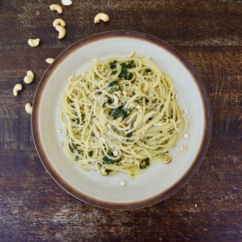 A plate of Thai basil pesto pasta topped with love oil and crushed cashew nuts surrounded by scattered cashews