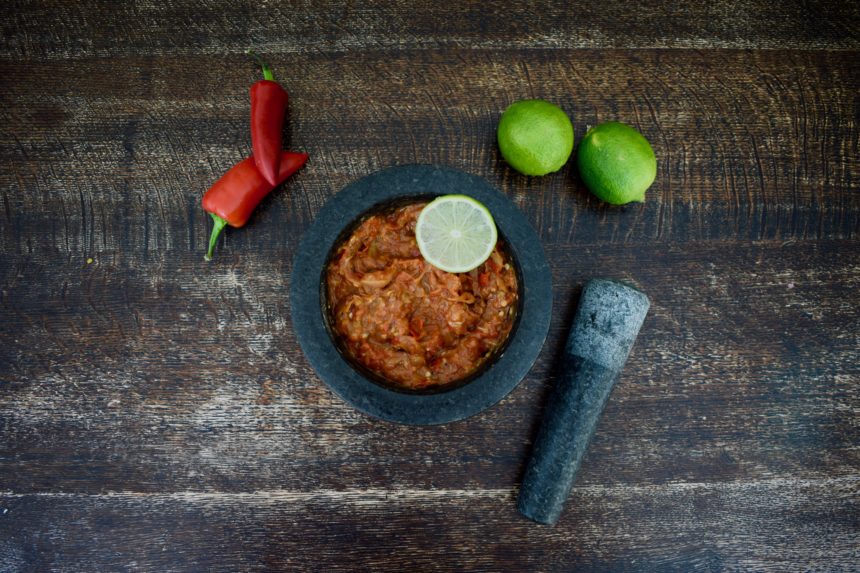 Sambal terasi Indonesia in a mortar on a wooden table next to a pestle, limes and raw red chillies