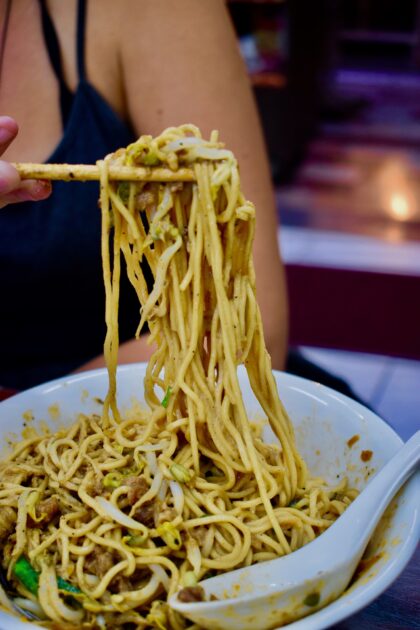 Picking up a large clump of sesame noodles from bowl with chopsticks