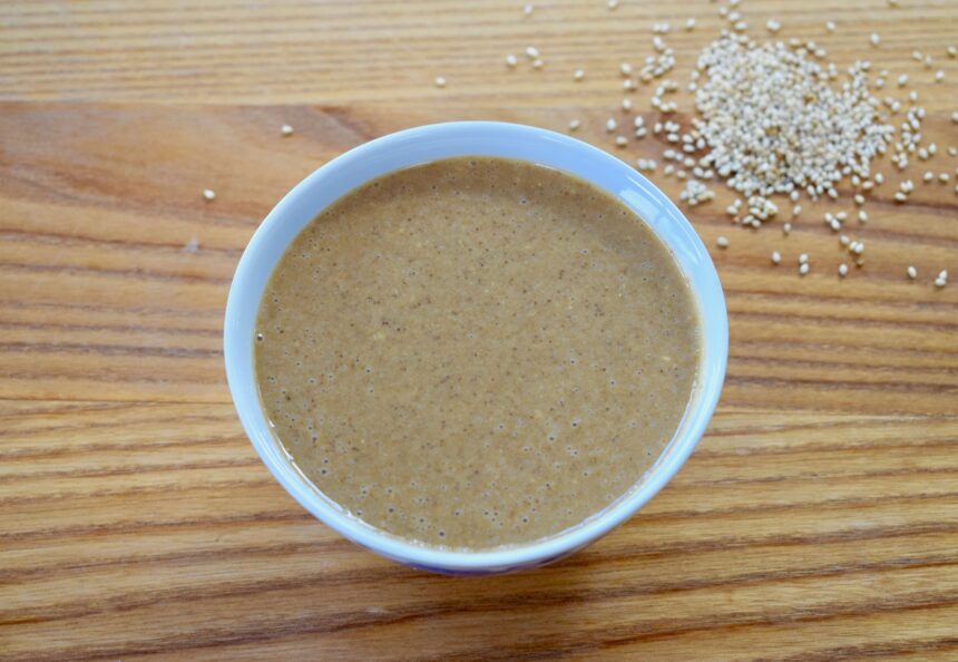 A bowl of homemade Chinese sesame paste (zhi ma jiang) with sesame seeds scattered on a wooden table