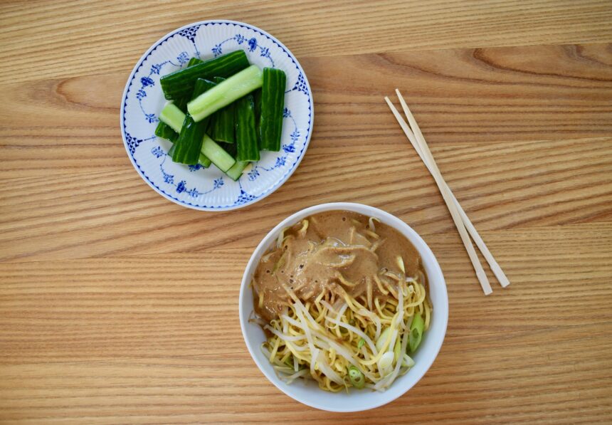 Bowl of Taiwanese cold noodles (liang mian) on a wooden table with chopsticks and bowl of Taiwanese cucumber salad