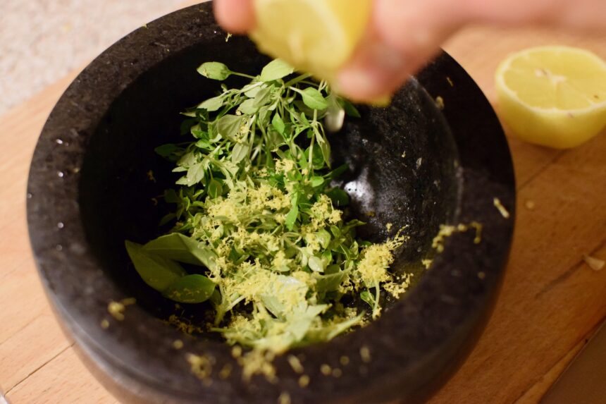 Hand squeezing a lemon into a pestle and mortar containing greek basil, lemon zest and garlic