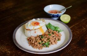 Thai basil minced pork (pad kra pao moo saap) with white rice and a fried egg on a table with lime and a small bowl of prik nam pla